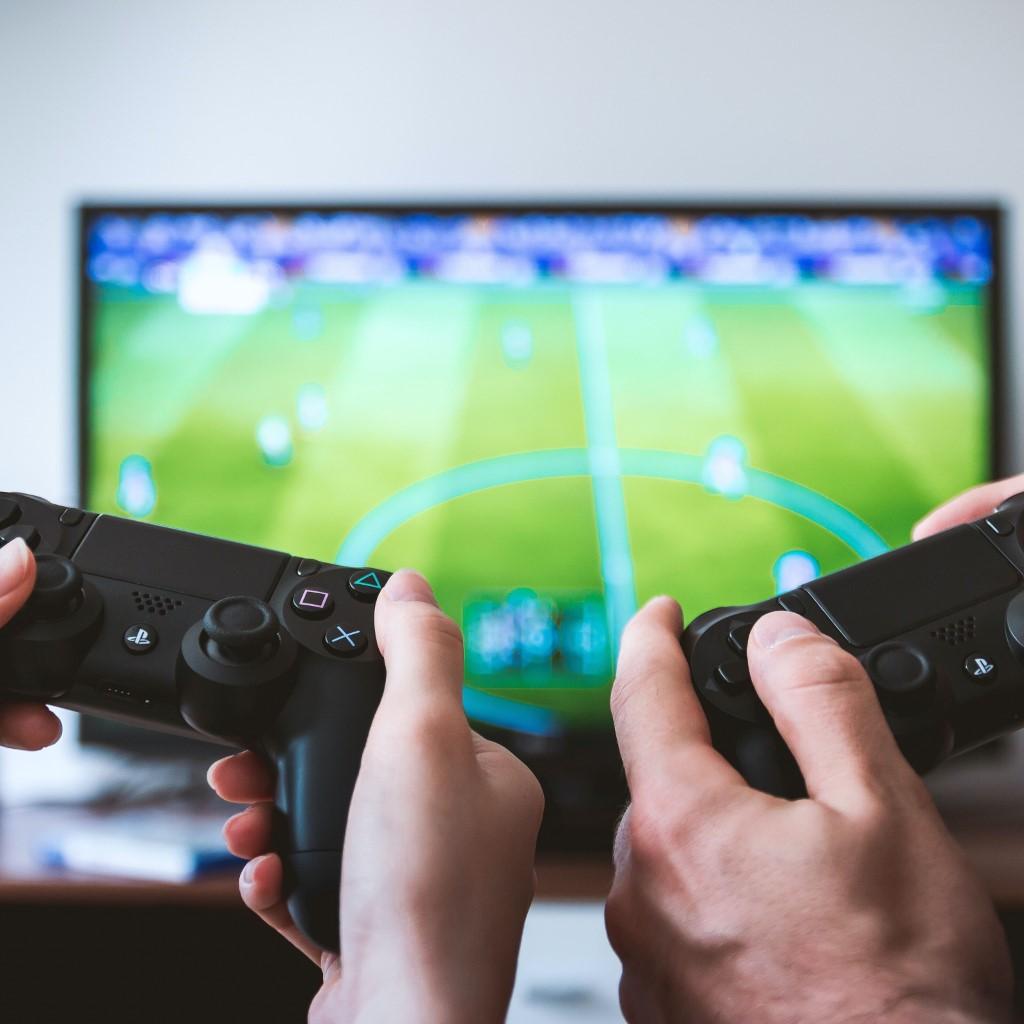 A close-up of two people's hands on controllers with a TV in the distance showing the video game being played.