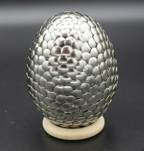 small egg covered in silver scales made from thumbtacks sitting on a light beige ring